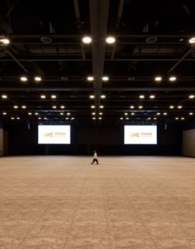 Inside a very large room in Tākina, 2 large projector screens hang in the background, a person stands in the middle. The brown floor and walls are minimal and the roof has many lights hanging down.