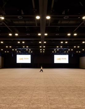 Inside a very large room in Tākina, 2 large projector screens hang in the background, a person stands in the middle. The brown floor and walls are minimal and the roof has many lights hanging down.