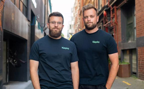 Two men in black tee shirts stand next to one another in an urban alleyway. 