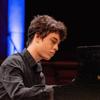 A young man wears a black collared shirt and plays a Steinway piano on stage.