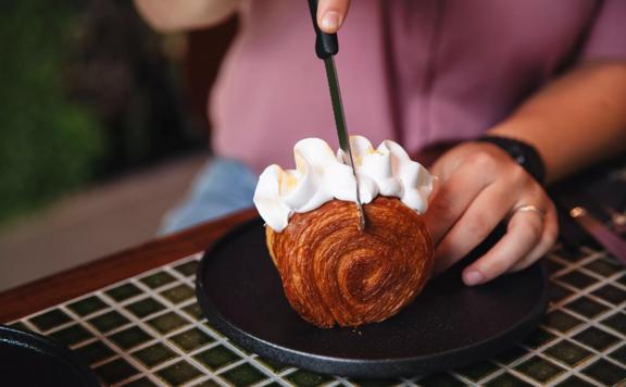 A person cuts a round pastry with a squiggle of cream on top. It sits on a black plate on a green tile table.