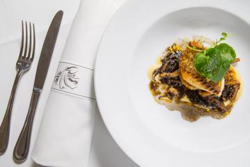 On a pristine white tablecloth sits silver cutlery, a napkin, and a white plate with grilled fish.