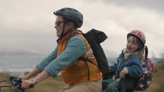 A production still from the series After the Party filmed in Wellington. Penny (Robyn Malcolm) rides a bicycle with Walt (played by Ziggy O'Reilly) in the rear seat with a body of water and a cloudy sky in the background. 