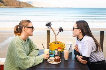 Two people enjoy drinks and a pastry on a patio table with a beach in the background.