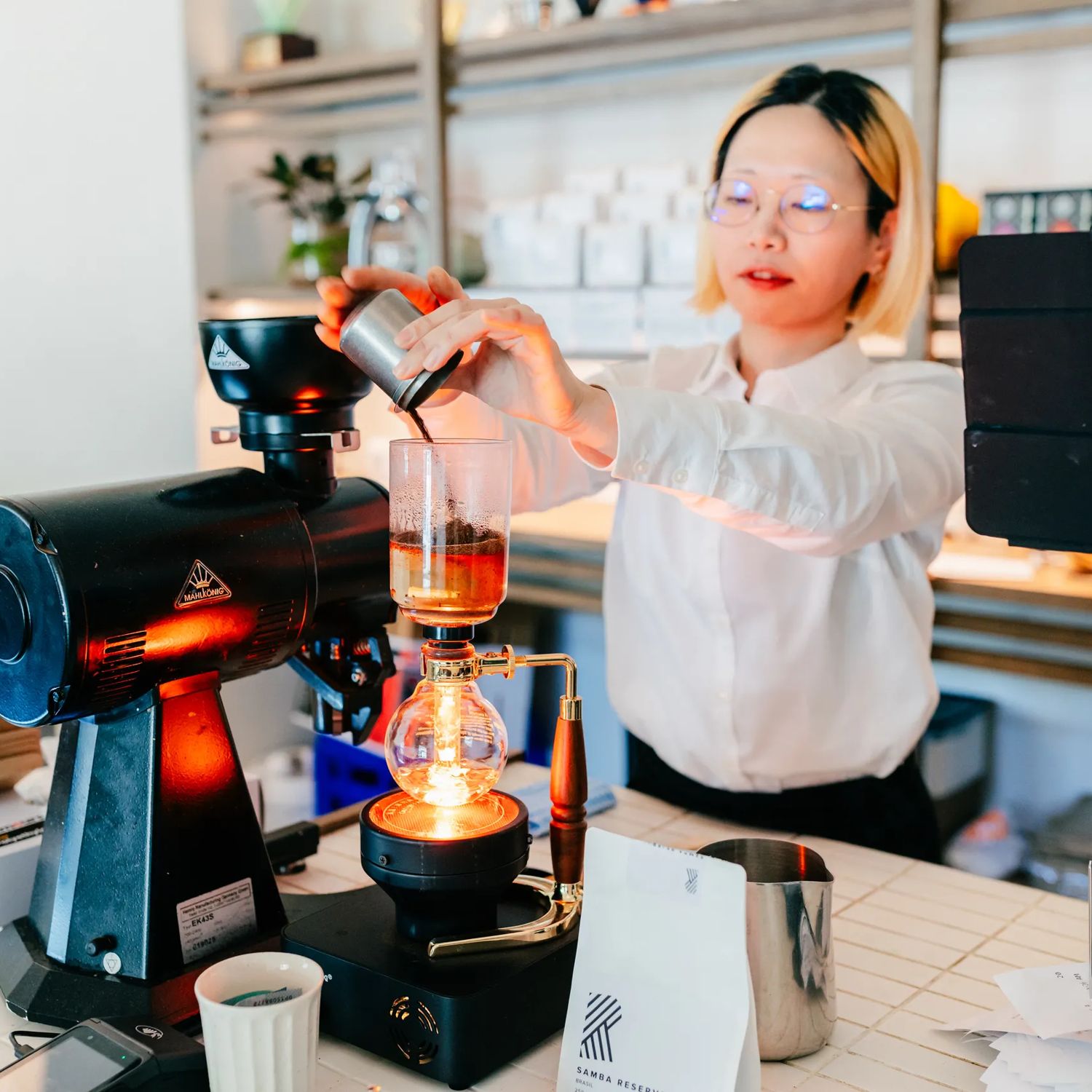 Barista preparing pour-over coffee with glass brewing equipment at a bright, modern café counter.