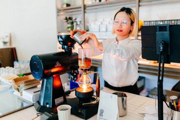 Barista preparing pour-over coffee with glass brewing equipment at a bright, modern café counter.