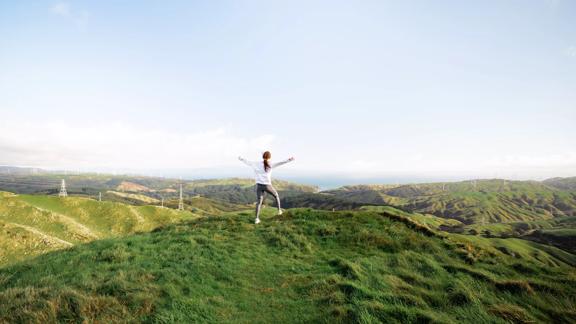 A person wears a white shirt and grey jogging pants is walking with their arms raised. They are facing the scenic vista in the background on the Skyline Walkway trail in Wellington. 