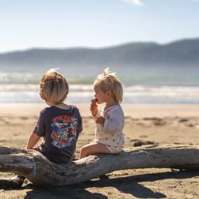2 young children sit on a log eating ice cream, on Paraparaumu Beach overlooking Kapiti island.