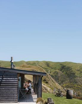 People sit in The Lodge wooden building at Boomrock. There are green hills behind and two chairs facing the ocean.