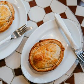 Two golden-brown pies on white plates with forks and folded napkins, arranged on a tiled café table at Jo's Pies in Brooklyn, Wellington.