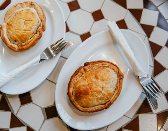Two golden-brown pies on white plates with forks and folded napkins, arranged on a tiled café table at Jo's Pies in Brooklyn, Wellington.