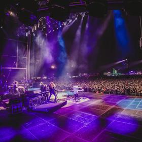 Musical group Toto performs on stage in purple spotlights in front of a packed audience.