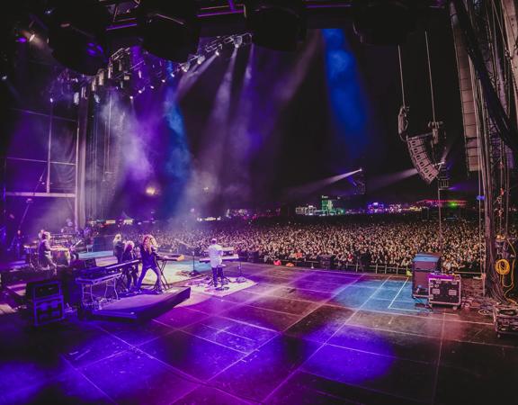 Musical group Toto performs on stage in purple spotlights in front of a packed audience.