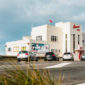 Exterior shot of Maranui cafe, on the top floor of the Lyall Bay surf life-saving club. The building is white with red accents and cars are parked outside.