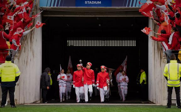 A still from the film Red White and Brass of a marching band entering a stadium. They are wearing red and white uniforms and the audience is cheering wearing red and white and waving the Tongan flag.