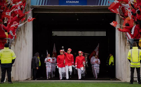 A still from the film Red White and Brass of a marching band entering a stadium. They are wearing red and white uniforms and the audience is cheering wearing red and white and waving the Tongan flag. 