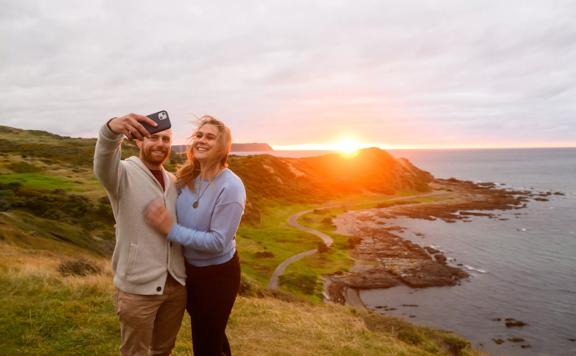 Two people take a selfie on a beautiful coastline in front of the sunset at Whitireia Park in Porirua, New Zealand.