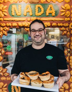 A baker stands outside Nada Bakery holding a tray of savoury pies, with the bakery’s decorative storefront and signage in the background.