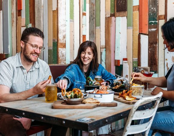 Three people eat crab and other seafood plates at The Crab Shack on Queens Wharf.