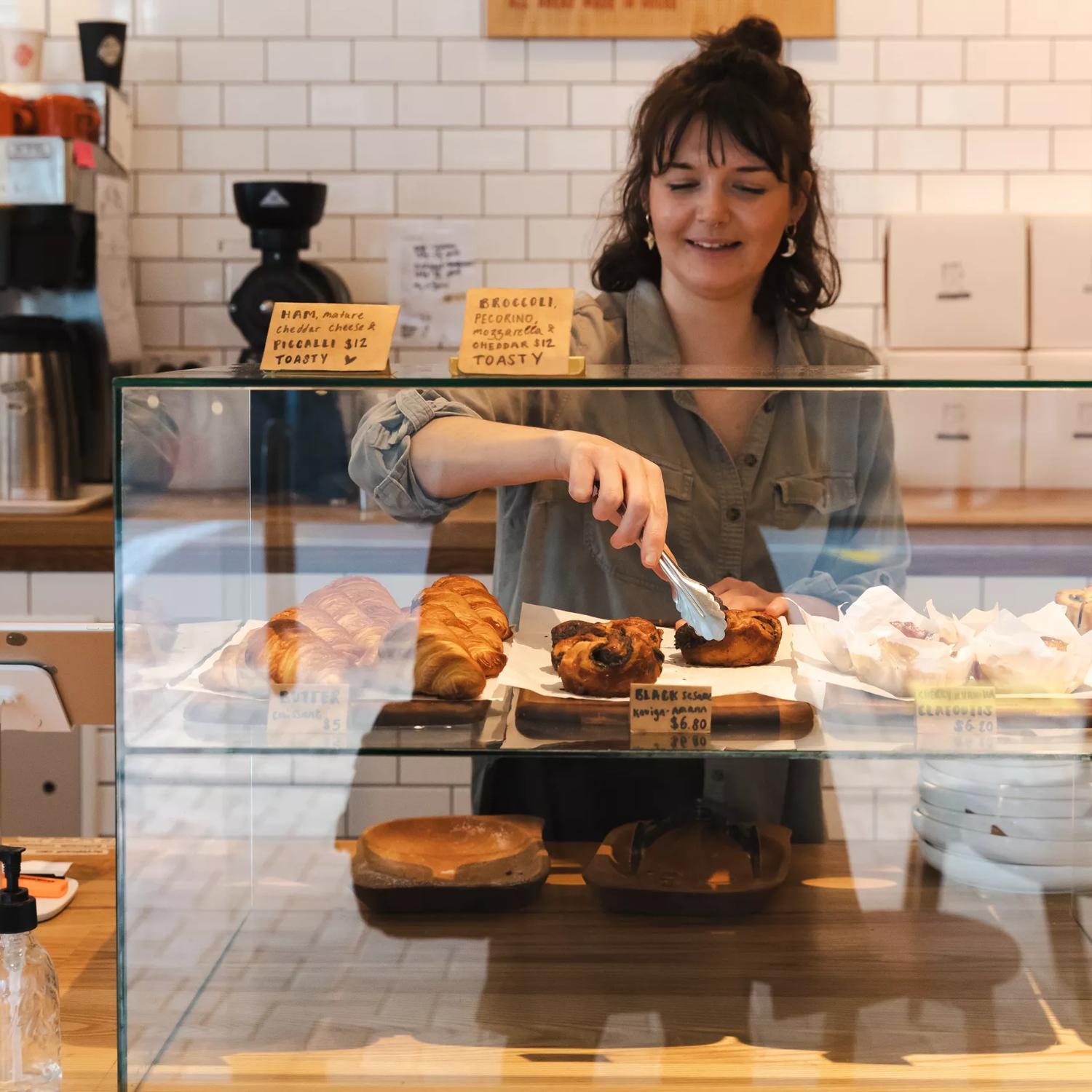 The front counter at Little Grump bakery located in Wellington Central. The back wall is covered in a white subway tile, the counter is a light-coloured wood and a worker is using tongs to pick up a pastry.