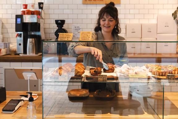 The front counter at Little Grump bakery located in Wellington Central. The back wall is covered in a white subway tile, the counter is a light-coloured wood and a worker is using tongs to pick up a pastry.