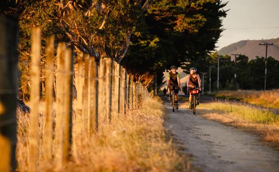 2 bikers cycling along the track next to the road, on the Western lake Road Section of the Remutaka Cycle Trail.