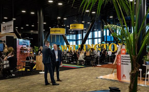 At MEETINGS 2023 at Tākina two people stand in the middle of the venue surrounded by exhibition booths.