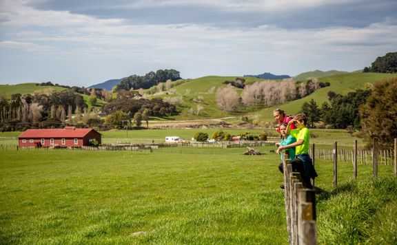 Three people in neon clothing leaning against a wooden farm fence, looking out at the view of farmland and a red farm building.