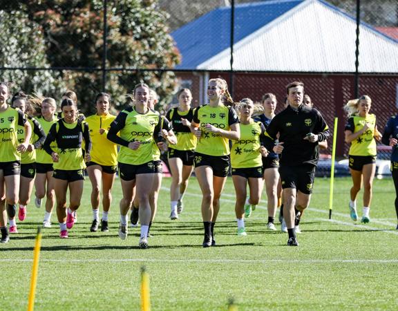 Wellington Phoenix women football players are dressed in yellow shirts and running as a group on a grass pitch.