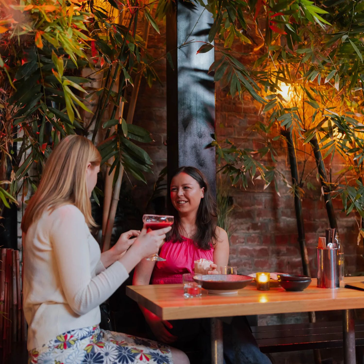 Two friends enjoying cocktails in a courtyard surrounded by bamboo plants.