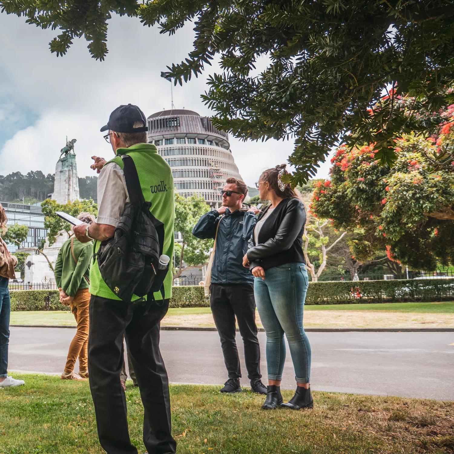 A group of tourists on a walking tour of Wellington, New Zealand. The guide wears a lime green vest and gestures toward the Beehive,  the Executive Wing of NZ Parliament Buildings, located at the corner of Molesworth Street and Lambton Quay.