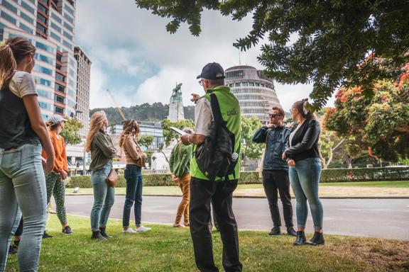 A group of tourists on a walking tour of Wellington, New Zealand. The guide wears a lime green vest and gestures toward the Beehive,  the Executive Wing of NZ Parliament Buildings, located at the corner of Molesworth Street and Lambton Quay.