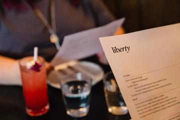 The corner of a menu from Liberty, with two water glasses, a pink cocktail in a collins glass and a person holding another menu, is out of focus in the background.