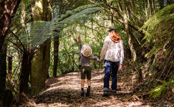 A section of trail Te Ara o Tama in Ōtari-Wiltons Bush. A parent and child hold hands and walk along the path as the child points upward.