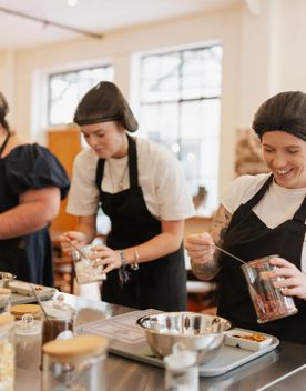 Three people on the Chocolatier experience at Wellington Chocolate Factory. They are all scooping toppings from a jar onto chocolate.