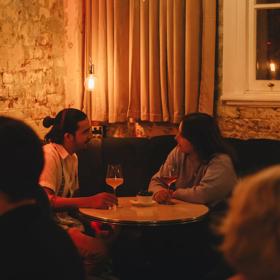 Two people converse over a glass of wine in the corner booth at Puffin wine bar, a natural wine and cocktail bar in Te Aro, Wellington. 