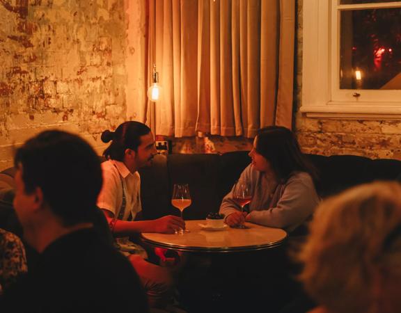 Two people converse over a glass of wine in the corner booth at Puffin wine bar, a natural wine and cocktail bar in Te Aro, Wellington.