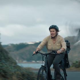 A production still from the series After the Party filmed in Wellington. Penny (played by Robyn Malcolm), wearing a helmet and a beige blouse, rides a bicycle on a road with a blurry seagull taking flight in the foreground. 