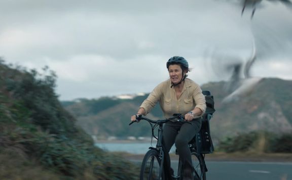 A production still from the series After the Party filmed in Wellington. Penny (played by Robyn Malcolm), wearing a helmet and a beige blouse, rides a bicycle on a road with a blurry seagull taking flight in the foreground.