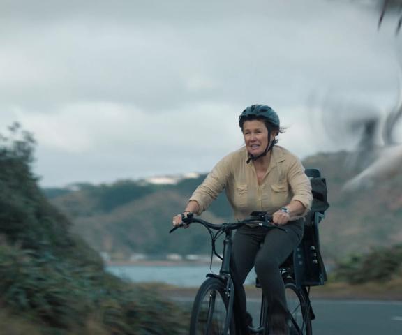 A production still from the series After the Party filmed in Wellington. Penny (played by Robyn Malcolm), wearing a helmet and a beige blouse, rides a bicycle on a road with a blurry seagull taking flight in the foreground. 