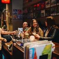 People seated at a bar, holding drinks and talking with a bartender, with records displayed on the wall behind.