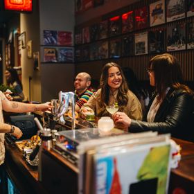 People seated at a bar, holding drinks and talking with a bartender, with records displayed on the wall behind.