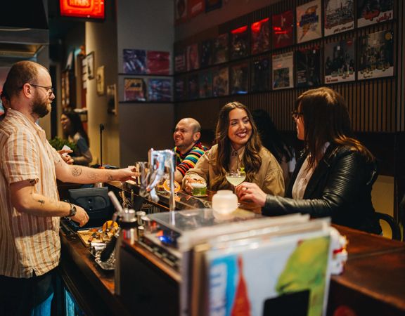 People seated at a bar, holding drinks and talking with a bartender, with records displayed on the wall behind.
