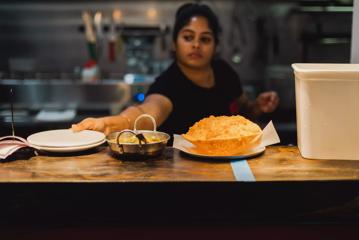 A chef at Chaat Street is working in the kitchen. They are placing a curry dish in the pass-through window.