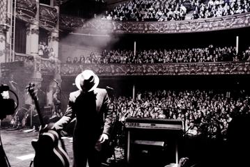 A musician wearing a fedora and a suit is on stage with his back to a packed cheering audience and puts down an acoustic guitar.