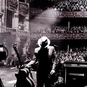 A musician wearing a fedora and a suit is on stage with his back to a packed cheering audience and puts down an acoustic guitar.