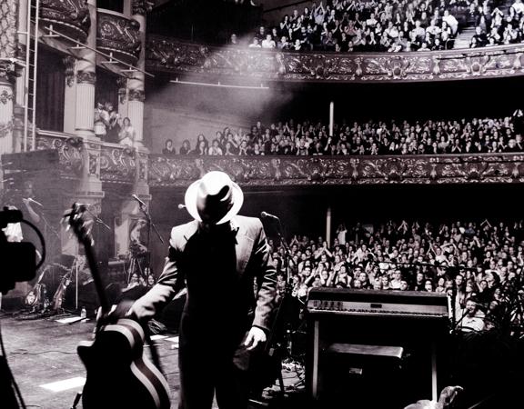 A musician wearing a fedora and a suit is on stage with his back to a packed cheering audience and puts down an acoustic guitar.