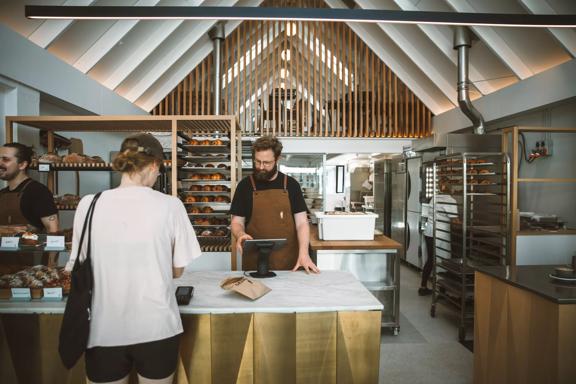 A person is paying for their order at the counter of Baker Gramercy, a bakery in Upper Hutt. The interior has a modern, minimalist feel with white walls and light wooden accents. 
