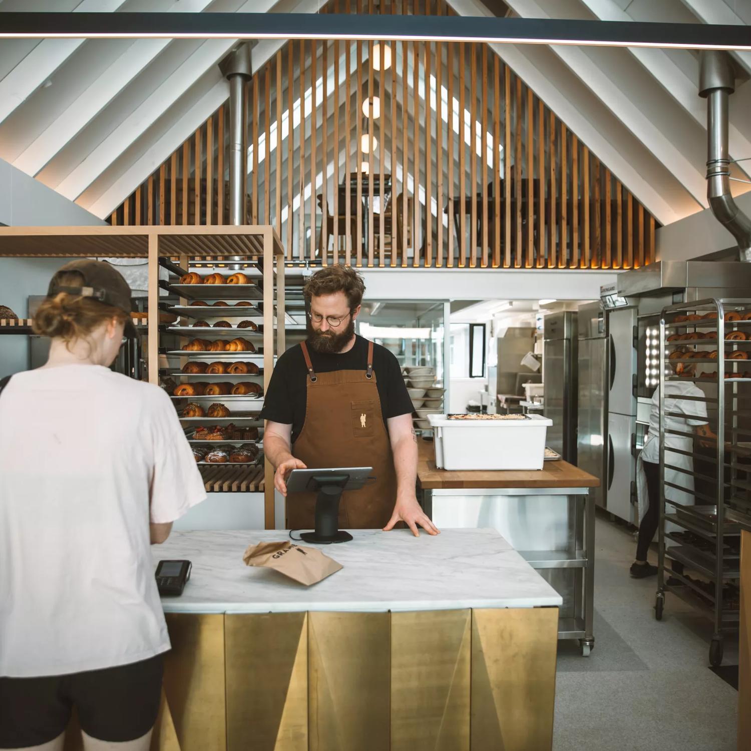 A person is paying for their order at the counter of Baker Gramercy, a bakery in Upper Hutt. The interior has a modern, minimalist feel with white walls and light wooden accents.