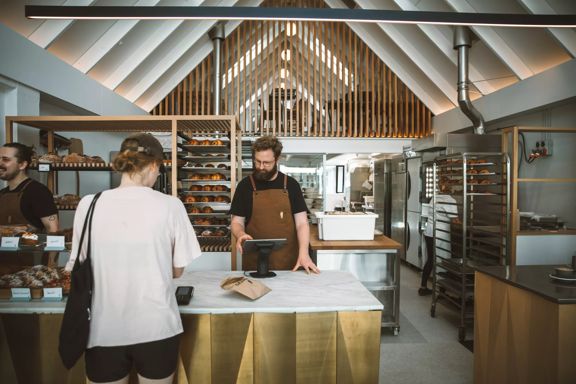 A person is paying for their order at the counter of Baker Gramercy, a bakery in Upper Hutt. The interior has a modern, minimalist feel with white walls and light wooden accents.